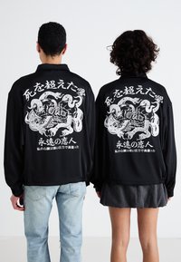 Črne majice z dolgimi rokavi imajo zapletene bele grafične vzorce, vključno s lobanjami in kačami, ter japonskim besedilom na hrbtu.