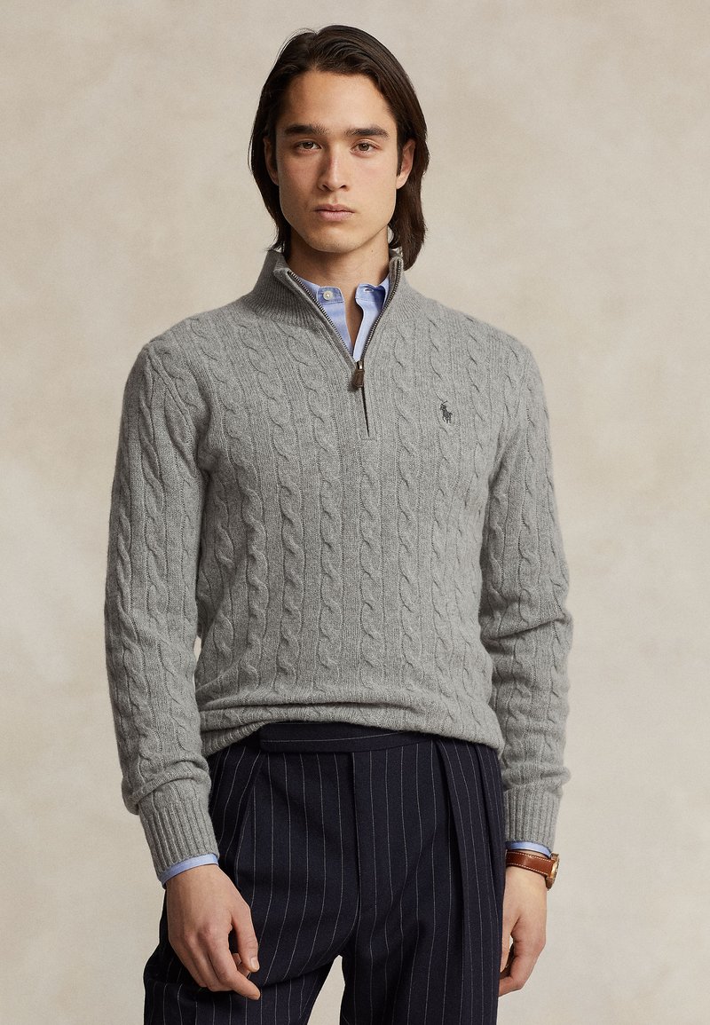 Uomo che indossa un maglione grigio chiaro a trecce con zip fino a un quarto sopra una camicia blu, pantaloni scuri a righe sottili e un orologio da polso marrone.