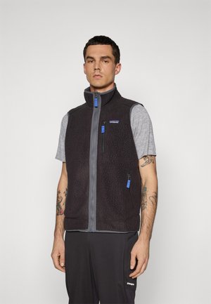 Man wearing black fleece vest over gray short-sleeve shirt, with visible arm tattoos, standing against plain light background.