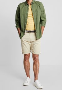 Camicia button-up verde in lino sopra una maglietta gialla, abbinata a pantaloni corti beige arrotolati e sneakers bianche. Include una cintura a righe.