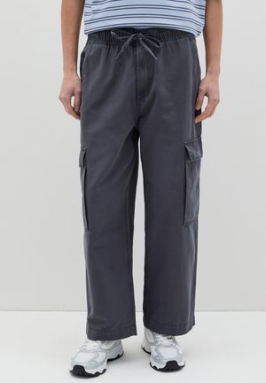 Pantalones cargo anchos y grises con cintura ajustable, bolsillos laterales y una textura suave, combinados con zapatillas blancas.
