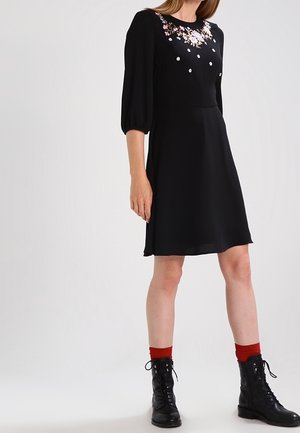 Robe noire avec broderies florales et accents blancs sur le corsage, manches bouffantes longues et jupe évasée. Portée avec des bottes noires et des chaussettes rouges.