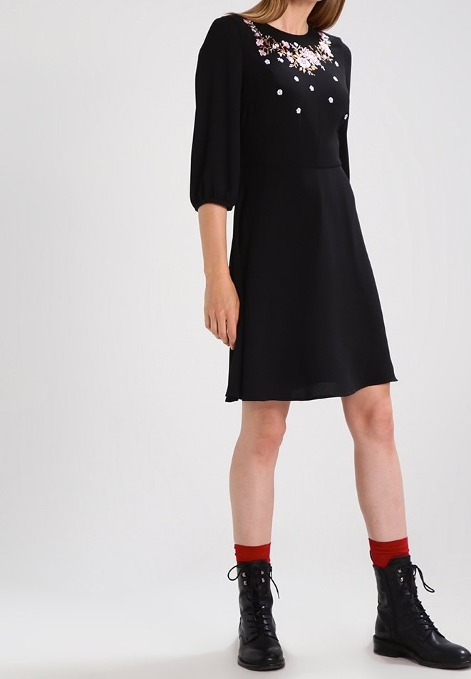 Robe noire avec broderies florales et accents blancs sur le corsage, manches bouffantes longues et jupe évasée. Portée avec des bottes noires et des chaussettes rouges.