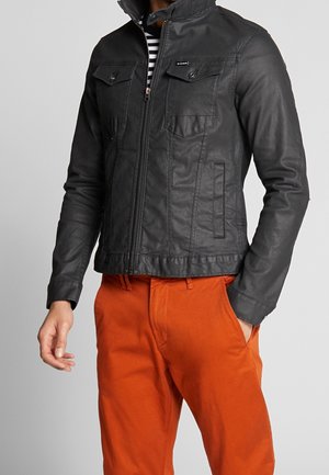 Mężczyzna w czarnej skórzanej kurtce z kieszeniami na piersi i pomarańczowych spodniach, jedna ręka w kieszeni, na jednolitym szarym tle.
