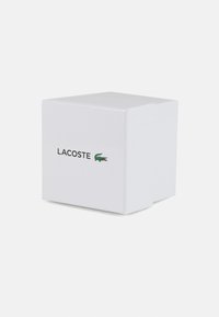 Caja cuadrada blanca con un acabado suave. Presenta un logo negro de "LACOSTE" y un ícono de cocodrilo verde en un lado.
