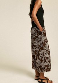 Pantalons larges marron avec un motif paisley blanc, fabriqués en tissu léger, associés à un haut sans manches noir et des sandales.