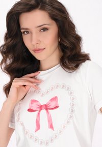 Hvid T-shirt med et pink buebånd design i et hjerte mønster lavet af små, lyse perler. Korte ærmer og rund halsudskæring.