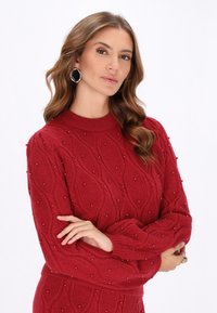 Czerwony sweter z dzianiny z teksturowanym wzorem liści i zdobieniami. Okrągły dekolt i długie rękawy. Modelka w koordynującym zestawie.