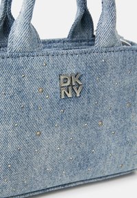 Denim handtas met kleine zilveren studs, voorzien van twee korte handvatten en een prominente zilveren DKNY-logo aan de voorkant. Lichtblauwe kleur.