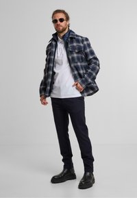 Giacca a quadri blu e bianca, sovrapposta a un polo bianco. Pantaloni neri slim-fit e stivali neri massicci completano l'outfit.