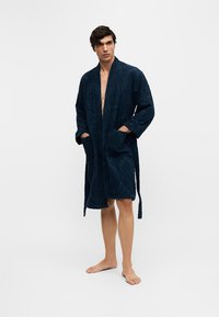 Jeune homme portant un peignoir bleu foncé à motifs, debout pieds nus les mains dans les poches devant un fond blanc uni.