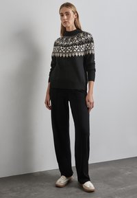 Schwarzer Pullover mit einem geometrischen cremefarbenen Muster am Ausschnitt, kombiniert mit schwarzen Hosen und cremefarbenen Sneakers mit einer strukturierten Sohle.