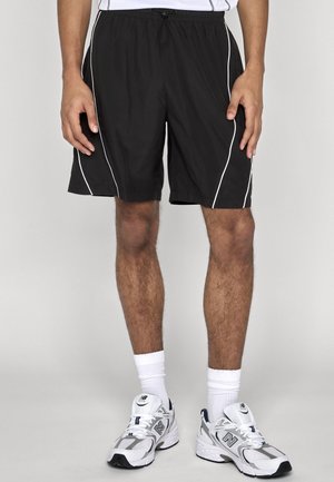 Mężczyzna w czarnych sportowych szortach z białym obszyciem, białych skarpetkach i białych sneakersach New Balance, stojący na jednolitym tle.