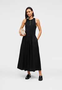 Μαύρο φόρεμα χωρίς μανίκια με εφαρμοστό μπούστο, φαρδύ φόρεμα και τρία χρυσά κουμπιά στο λαιμό. Λεπτή υφή· φορεμένο με μαύρα λοφάρ.