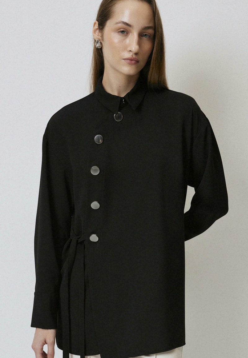 Zwarte blouse met een kraag, knoopsluiting aan de voorkant, een zijstrikdetail en lange mouwen. Gemaakt van een zachte, lichte stof.