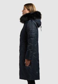 Parka blu scuro con cappuccio foderato in pelliccia sintetica, chiusura con zip e bottoni a pressione, tasche laterali e motivo trapuntato sulle maniche. Design lungo e aderente.