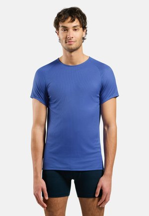 Mann steht mit Blick nach vorn, trägt ein blaues kurzärmeliges Hemd und schwarze enganliegende Shorts vor einem schlichten weißen Hintergrund.