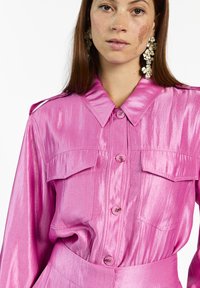Vrouw in een glanzende, felroze, knoopshirt met borstzakken en grote bloemenoorbellen tegen een witte achtergrond.