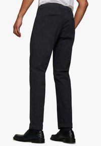 Pantalones azul marino oscuro con una tela texturizada, que presentan un corte ajustado y puños enrollados. Se pueden ver los bolsillos traseros, combinados con zapatos negros brillantes.
