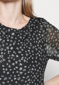 Närbild av en kvinna som bär en svart genomskinlig blommig blus med små vita blommönster och ett tunt guldhalsband.