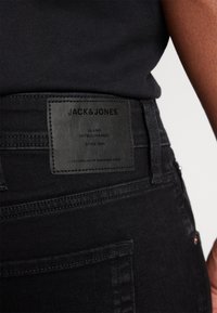 Pantalones vaqueros de mezclilla negra con una etiqueta de cuero texturizado que presenta "JACK&JONES" y "JEANS INTELLIGENCE" en relieve negro. Diseño clásico de corte recto.