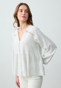 Biała bluzka z długim rękawem w pionowe paski, z dekoltem w serek, z zapięciem na guziki, w stylu peplum, z koronkowymi akcentami na ramionach. Miękka faktura materiału.