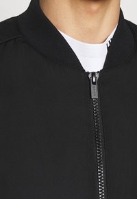 Giacca bomber nera con colletto a coste e chiusura centrale a cerniera. Presenta un tessuto liscio e un logo discreto sul cursore della cerniera.