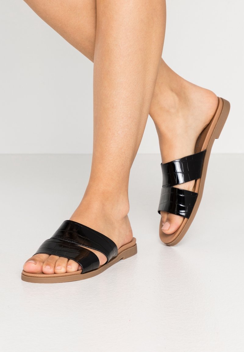 Pieds portant des sandales slip-on noires à deux brides avec texture crocodile et semelles plates couleur sable sur un sol clair.