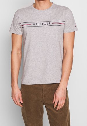 Mand iført en lysegrå T-shirt med "HILFIGER" og tynde sorte og røde striber hen over brystet, kombineret med brune kordbukser.