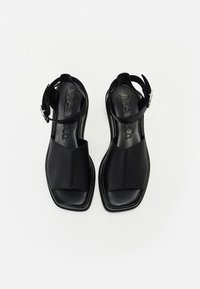 Czarne skórzane sandały z płaską podeszwą, otwartym palcem, szerokim paskiem z przodu oraz regulowanym paskiem na kostkę z metalowym zapięciem. Gładka tekstura.