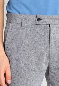 Šedé kalhoty na míru s texturovanou látkou, vybavené předním zapínáním na knoflík a bočními kapsami. Pásek má poutka na opasek.