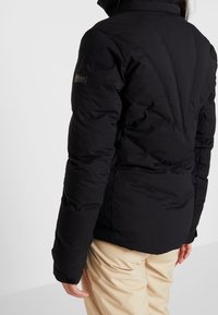 Doudoune noire avec un col montant, une texture matelassée et un logo sur la manche. Le modèle porte un pantalon beige.