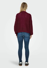 Burgundské pletené svetr s volným střihem, spadlými rameny a žebrovaným lemem, v kombinaci s tmavě modrými skinny džínami a černobílými teniskami.