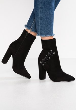 Černé semišové kotníkové boty s špičatou špičkou a vysokým blokovým podpatkem. Mají boční zip a dekorativní šněrování se stříbrnými očky.