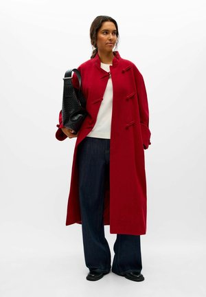 Une femme porte un long duffle-coat rouge, un haut blanc et un jean à jambes larges. Elle porte un sac d’épaule noir matelassé sur un fond blanc.