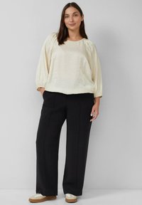 Blusa di colore crema, ampia e svasata, con maniche a sbuffo a tre quarti, abbinata a pantaloni neri a vita alta e sneakers beige.