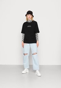 T-shirt oversized preta com a impressão "DEPARTAMENTO CRIATIVO", sobreposta a uma camisa de manga longa em padrão quadriculado preto e branco, combinada com jeans claros desgastados e botas brancas.