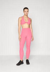Sutiã desportivo rosa com um design de frente torcida e leggings rosa de cintura alta. Ambos têm um acabamento suave. Sapatos desportivos pretos completam o visual.