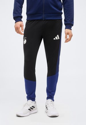 Homme portant un pantalon de survêtement noir et bleu avec des logos sportifs blancs et des chaussures de sport blanches sur un fond uni.
