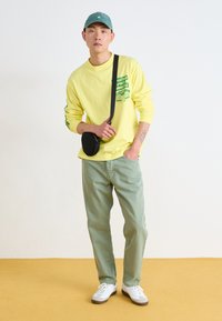 Camisa amarilla de manga larga con estampado verde, combinada con pantalones verde claro y zapatos blancos. Se lleva un pequeño bolso negro cruzado.