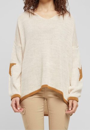 Femme portant un pull en maille crème avec des motifs d'étoiles marron sur les manches et un bord marron, associé à un pantalon beige avec poches latérales.