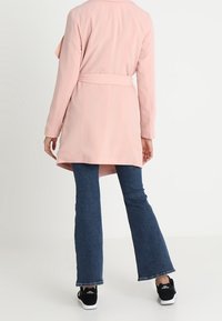 Cappotto rosa chiaro con vita cinturata, maniche lunghe e davanti drappeggiato. Abbinato a jeans denim a zampa e scarpe da ginnastica nere. Sfondo bianco semplice.