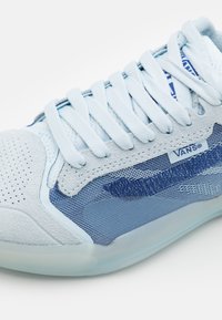 Sapatilha azul clara com um cabedal em camurça texturizada e malha, apresentando atacadores tonais, uma faixa azul e uma sola de borracha transparente.
