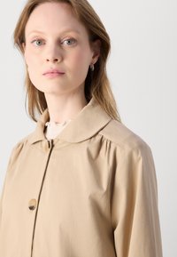 Besislė, lengva bežinė striukė su smailiu apykakle, mygtukų užsegimu ir susikaupusiomis peties detalėmis. Lygus paviršius ir minimalistiškas dizainas.