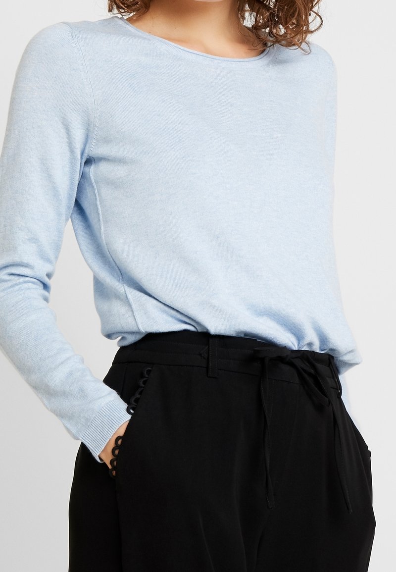 Pull à manches longues bleu clair en tissu tricoté doux, accompagné d'un pantalon noir taille haute avec ceinture à nouer et détails décoratifs sur les poches.