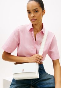 Λευκό δερμάτινο τσαντάκι ώμου με χρυσές λεπτομέρειες, κλείσιμο με καπάκι και αποσπώμενο λουρί, τοποθετημένο σε φωτεινό φόντο.