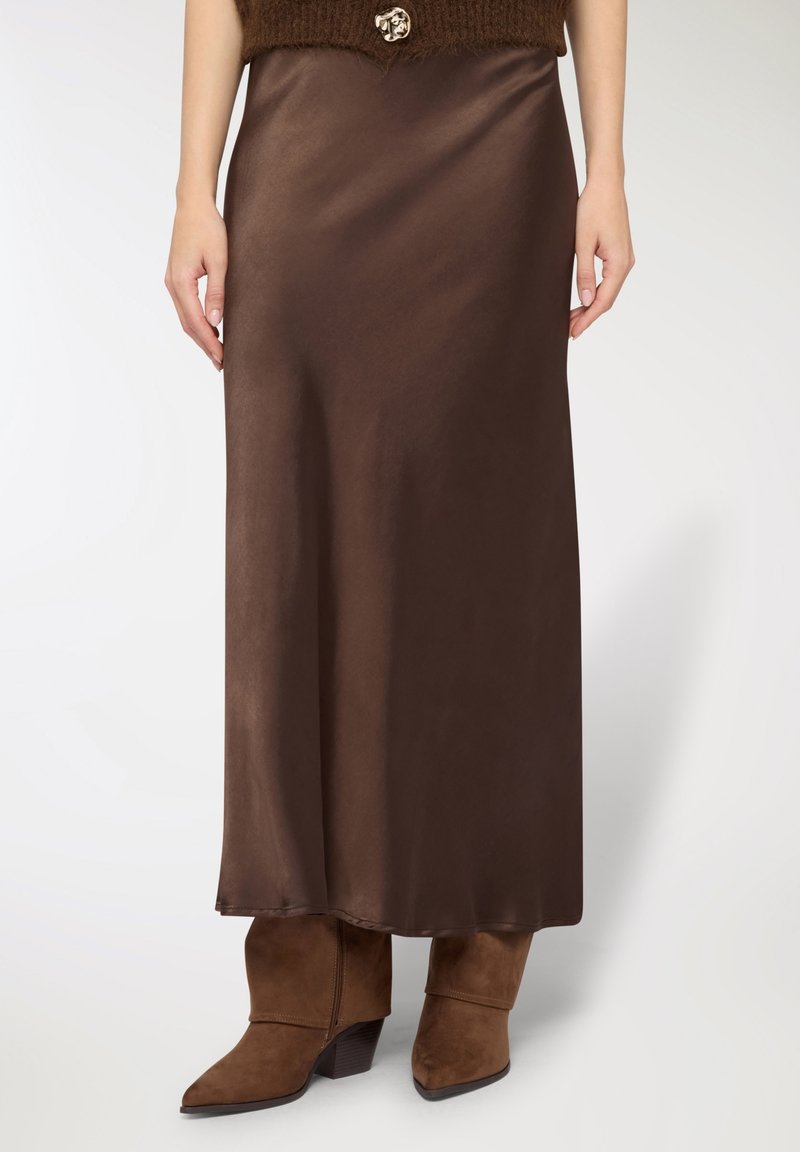 Gonna lunga in satin marrone con una texture liscia, abbinata a stivaletti color cuoio. L'outfit comprende un crop top marrone peloso con un bottone decorativo.