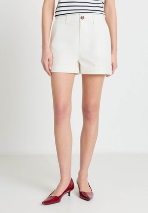 Femme portant un short blanc taille haute, un haut rayé et des chaussures rouges à talons bas et bouts pointus, debout sur un fond blanc uni.