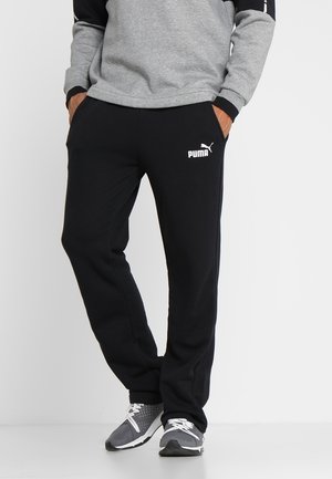 Czarne spodnie dresowe z bocznymi kieszeniami, elastyczną talią i białym logo Puma na lewej udzie. Noszone z szarymi butami sportowymi.