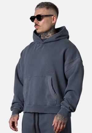 Mann mit Tattoos, der schwarze Sonnenbrille und einen dunkelgrauen übergroßen Hoodie mit Vordertasche trägt und seitlich vor einem schlichten Hintergrund schaut.